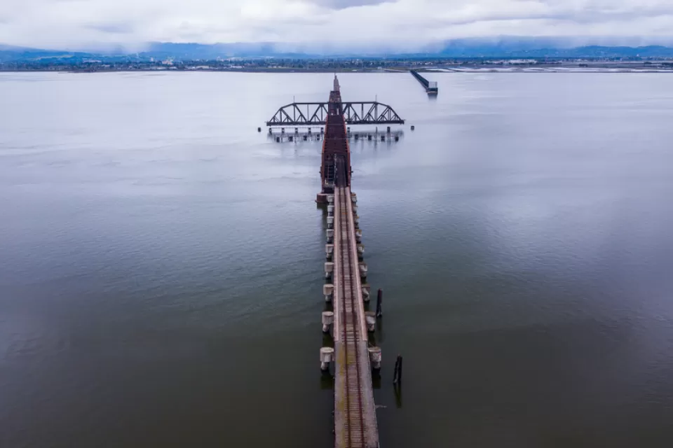 A long rail bridge, rusted and old, crosses a long stretch of water with hills and clouds on the other side.